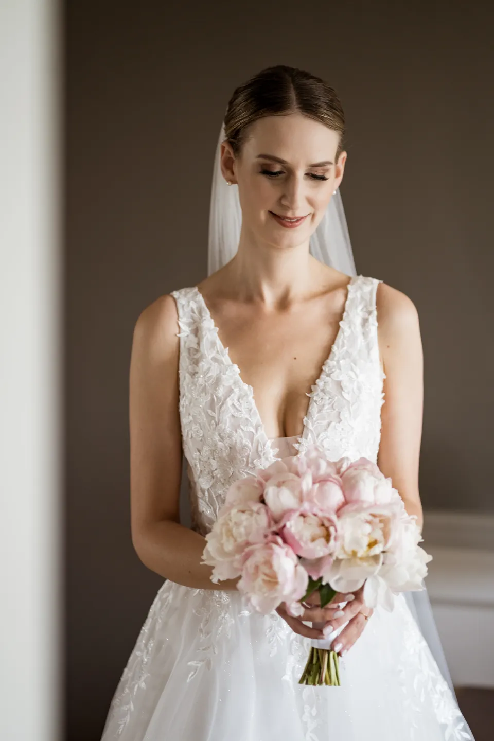 Braut im Spitzenkleid mit Schleier und Pfingstrosen-Strauß, natürliches Braut-Make-up und Hochsteckfrisur