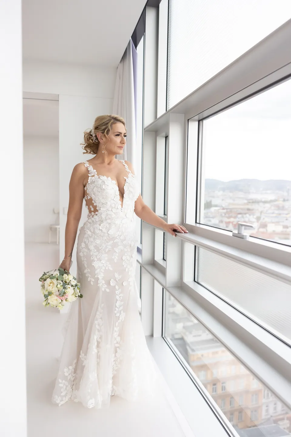 Braut im Spitzenkleid am Fenster mit Blick über die Stadt